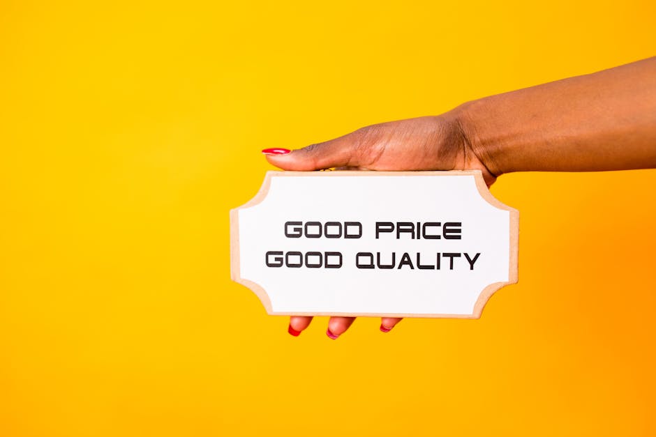A close-up of a person's hand with red nail polish holding a small, rectangular sign with a white background and a decorative wooden border, displaying the text 'Good Price Good Quality' in bold black letters. The background is a solid, bright yellow color, creating a vibrant contrast with the hand and sign. The image emphasizes affordability and quality, which are key considerations during home relocation or furniture transport services. This visual can be associated with professional removals, packing, and moving logistics offered by Man with Van Harefield, highlighting the importance of choosing reliable moving solutions.