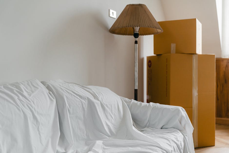 Inside a residential room, several cardboard moving boxes of various sizes are stacked against a light-colored wall. The boxes are sealed with packing tape and appear to be prepared for a home relocation service. To the left, there is a sofa covered with a white protective sheet, likely for furniture transport or packing during the moving process. Behind the boxes, a tall floor lamp with a brown pleated lampshade is positioned near the wall, indicating a typical living room environment. The room is illuminated by natural light coming from an unseen window. The setting suggests a final phase of packing or loading items into a moving vehicle, with visible furniture and packaging materials indicating an ongoing or preparatory stage of furniture transport, consistent with house removals and relocation services offered by Man with Van Harefield.