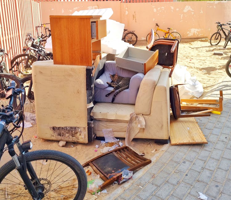 A pile of discarded household furniture and waste materials located on a paved outdoor area next to a row of parked bicycles, with a weathered brick and concrete wall in the background. The collection includes an old, stained fabric armchair with a white cushion featuring a lying tabby cat, a broken wooden chair, a small wooden bedside cabinet, a disassembled wooden coffee table, and various cardboard boxes and plastic packaging. Some items are partially wrapped in protective padding, while others are simply placed on the ground or against the wall. The scene appears to depict a commercial or residential waste clearance, likely part of a home relocation or furniture disposal process by Man with Van Harefield, in readiness for collection or disposal.