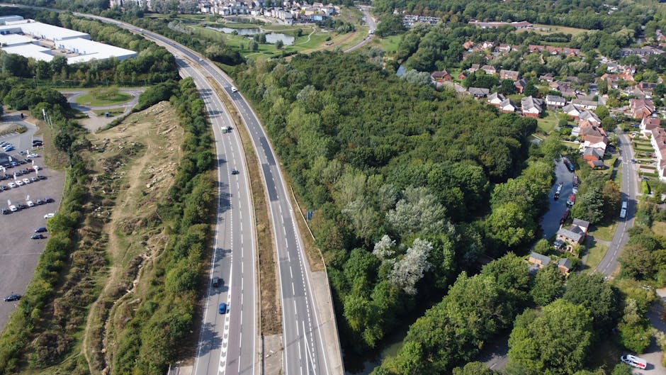 An aerial view of a dual carriageway with several vehicles driving along, bordered by dense green trees on one side and a mix of residential buildings with pitched roofs on the other. The road curves gently through a suburban area, with some cleared patches of earth and grassy areas on the left side near a parking lot. The scene is well-lit with natural daylight, illustrating the environment where home relocation and furniture transport logistics may take place, supported by professionals like Man with Van Harefield during their removals and loading processes.
