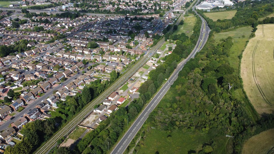 A narrow, rural road leading into a densely wooded area with tall, leafy green trees on both sides. The asphalt surface features white lane markings, including a small intersection or turning point in the foreground. The road appears to gently incline and disappear into the distance among the trees, with natural daylight illuminating the scene. This setting is representative of quiet, countryside pathways often encountered during home relocations or furniture transport in rural areas, aligning with services provided by Man with Van Harefield. The image emphasizes the importance of navigating narrow, tree-lined streets when planning a house move or loading process for furniture and packing materials, highlighting the types of environments that require careful logistical planning during a local move or removal service.