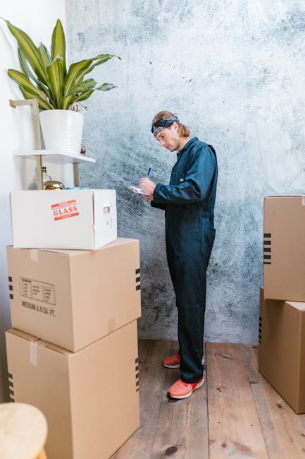 A man wearing a dark blue overalls and protective glasses is standing indoors against a textured light grey wall, surrounded by several packed cardboard boxes, some labeled and sealed with packing tape. The boxes are stacked on a wooden floor, with one partially visible on the left and others arranged around him, indicating a packing or home relocation process. He is holding a pen and appears to be making notes or inventory on a notepad, suggesting an organized approach to packing and moving logistics. To his left, a large potted plant with broad green leaves sits on a small white stand, adding a natural element to the scene. This setting captures the preparation stage of furniture transport and packing within a house removal or relocation service. The environment is well-lit, emphasizing the professional and methodical aspect of home moving services provided by Man with Van Harefield, fitting the context of emergency house removals and detailed load management.