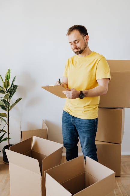 A man wearing a dark blue overalls and protective glasses is standing indoors against a textured light grey wall, surrounded by several packed cardboard boxes, some labeled and sealed with packing tape. The boxes are stacked on a wooden floor, with one partially visible on the left and others arranged around him, indicating a packing or home relocation process. He is holding a pen and appears to be making notes or inventory on a notepad, suggesting an organized approach to packing and moving logistics. To his left, a large potted plant with broad green leaves sits on a small white stand, adding a natural element to the scene. This setting captures the preparation stage of furniture transport and packing within a house removal or relocation service. The environment is well-lit, emphasizing the professional and methodical aspect of home moving services provided by Man with Van Harefield, fitting the context of emergency house removals and detailed load management.