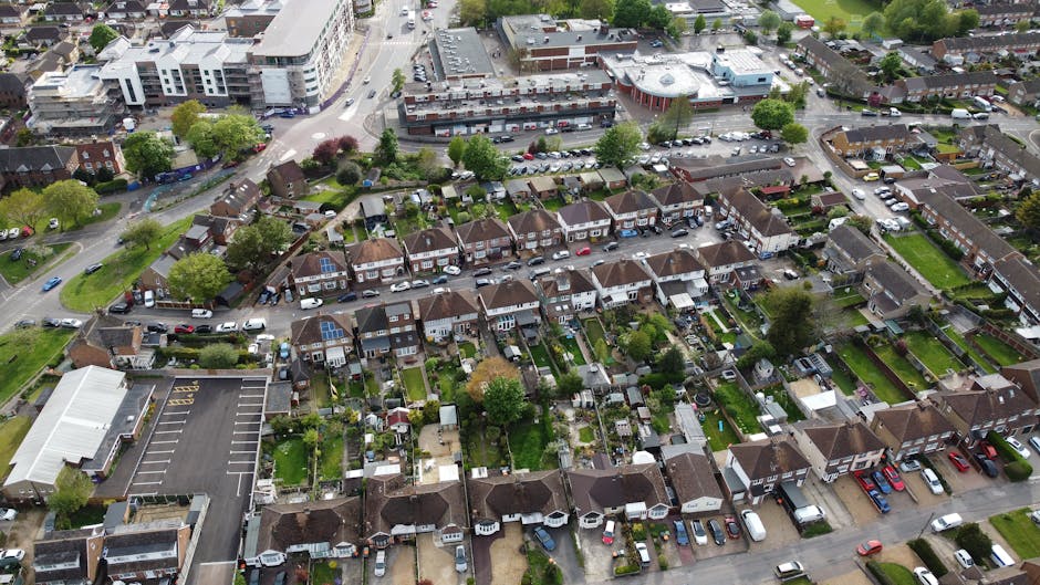 An aerial view of a residential street during daytime showing numerous semi-detached and terraced houses with small gardens, driveways, and parked cars. The foreground features a suburban neighbourhood with houses fitted with solar panels on their roofs, green lawns, and backyard patios with garden furniture and sheds. A large commercial building with a flat roof and parking lot is visible at the bottom left corner. To the back, a wider industrial or office complex with multiple floors and a parking lot with parked vehicles can be seen. The area is surrounded by trees and greenery. This image captures the infrastructure typical of a home relocation area, emphasizing the property's access points and nearby transport routes, relevant to house removals and furniture transport services like those provided by Man with Van Harefield, as referenced in the context of applying for Hillingdon Council permits for removal vans, which are often used during packing, moving, and loading processes.