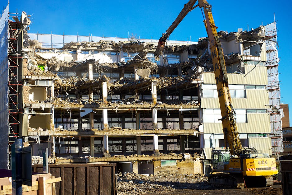 A multi-storey building is in the process of being demolished, with large sections of walls and floors partially torn away, exposing the internal structure and spaces. The building's facade shows broken panels, debris, and exposed concrete, with some sections leaning or missing. A yellow demolition excavator with a long arm and bucket attachment is actively working on the upper floors, positioned on a clear area of ground in front of the building. Scaffolding surrounds parts of the structure, and safety netting is visible on the sides. The construction site is outdoors, under a bright blue sky, with sunlight highlighting the remaining elements of the building and the machinery. This demolition scene reflects the ongoing process of clearing space, which could be related to future property development or preparation for a new build, aligning with the themes of property management and construction services associated with house removals and building projects.