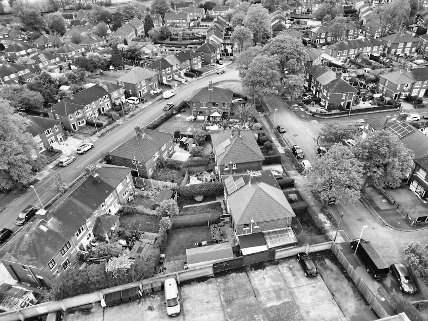 An aerial view of a suburban residential area showing multiple houses with pitched roofs, some with solar panels, and neatly maintained gardens enclosed by low fences. Several cars are parked along the street and in driveways. In the foreground, a property with a small backyard features a garden shed, patio area, and garden furniture. The street curves gently through the neighbourhood, with mature trees providing shade and greenery. The scene appears to be taken during daylight hours, with clear visibility of the various properties, pathways, and parked vehicles. The image captures a typical urban environment suitable for house removals or furniture transport services, with visible surroundings indicative of a quiet, established residential community in Hillingdon, suitable for the services provided by Man with Van Harefield related to packing, loading, and home relocation.