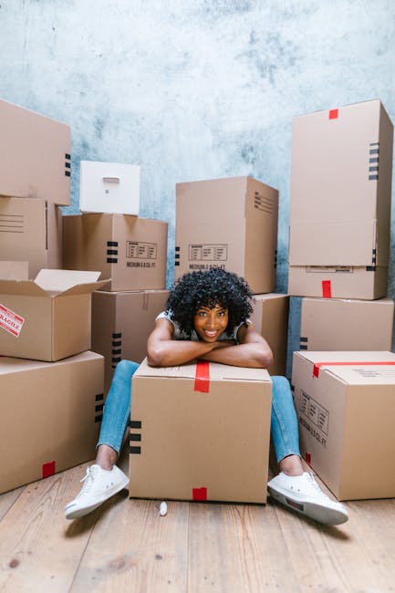 A young woman with curly black hair, wearing a white T-shirt, blue jeans, and white sneakers, is sitting cross-legged on a light wooden floor surrounded by numerous cardboard moving boxes of various s