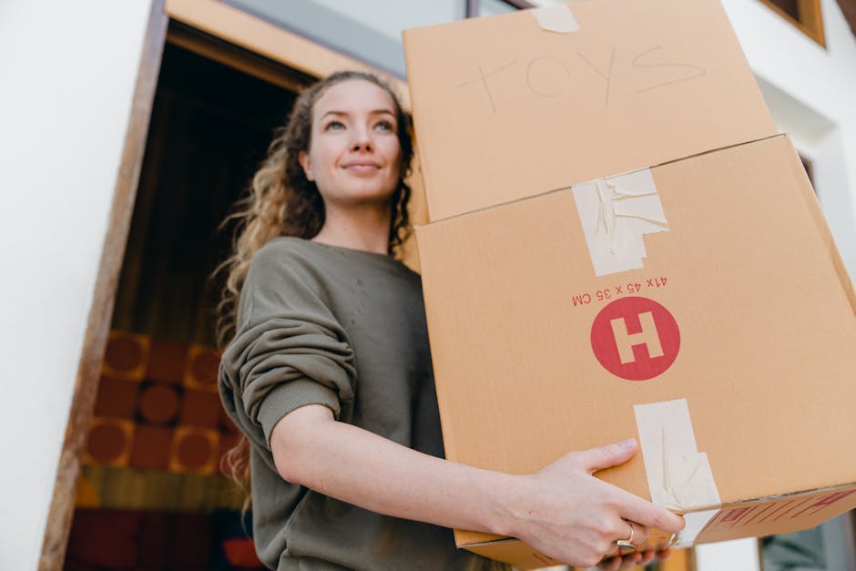 A young woman with curly black hair, wearing a white T-shirt, blue jeans, and white sneakers, is sitting cross-legged on a light wooden floor surrounded by numerous cardboard moving boxes of various s