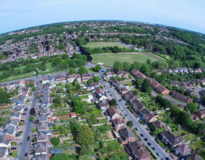 An aerial view of a suburban residential area with rows of terraced and semi-detached houses, tree-lined streets, and neatly maintained gardens. In the foreground, a street is visible with parked cars on both sides and houses with pitched roofs, some featuring small front yards. Behind this, there are larger green spaces, including a sports field or park, surrounded by trees, and additional residential streets extending into a densely built neighborhood. The scene is illuminated by natural daylight, with clear skies overhead, and the overall environment depicts a typical UK suburban community. This image is relevant to house removals and home relocation services provided by Man with Van Harefield, illustrating the type of area where professional moving and furniture transport operations are often carried out.