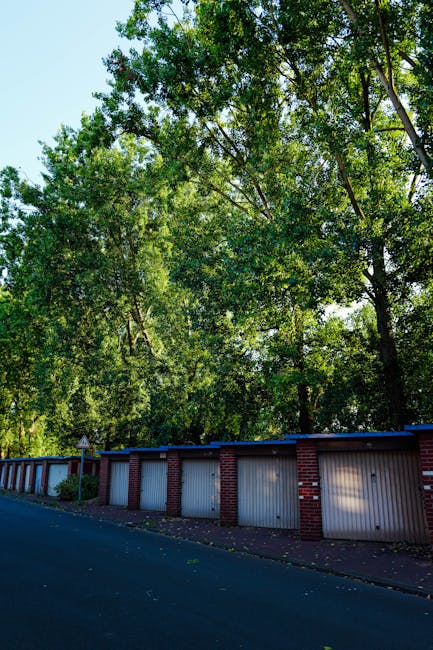 Exterior view of a row of closed garage units with white metal doors and red brick framing, situated along a paved street. Behind the garages, there is a dense canopy of tall, leafy trees with green f