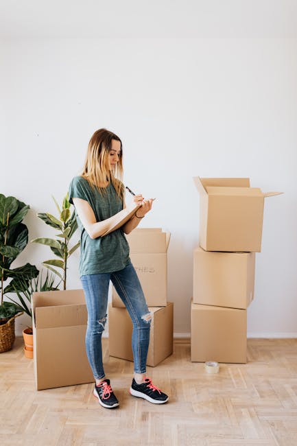 A young woman with shoulder-length blonde hair, dressed in a casual green t-shirt, ripped blue jeans, and black and pink sneakers, standing indoors on a wooden floor next to several packed cardboard moving boxes. She is writing on a clipboard or notepad, likely making a checklist related to her house move. Behind her, a tall green houseplant in a woven basket adds a natural element to the scene. One of the boxes is open with flaps folded outward, and another box is partially visible behind her. The setting suggests an interior space prepared for a home relocation, with visible packing materials and a focus on organizing belongings efficiently. The walls are plain white, creating a neutral backdrop suitable for a moving-related process, with natural lighting illuminating the scene. This image aligns with the activities involved in packing and moving, as part of a house removal or furniture transport service, such as those offered by Man with Van Harefield.