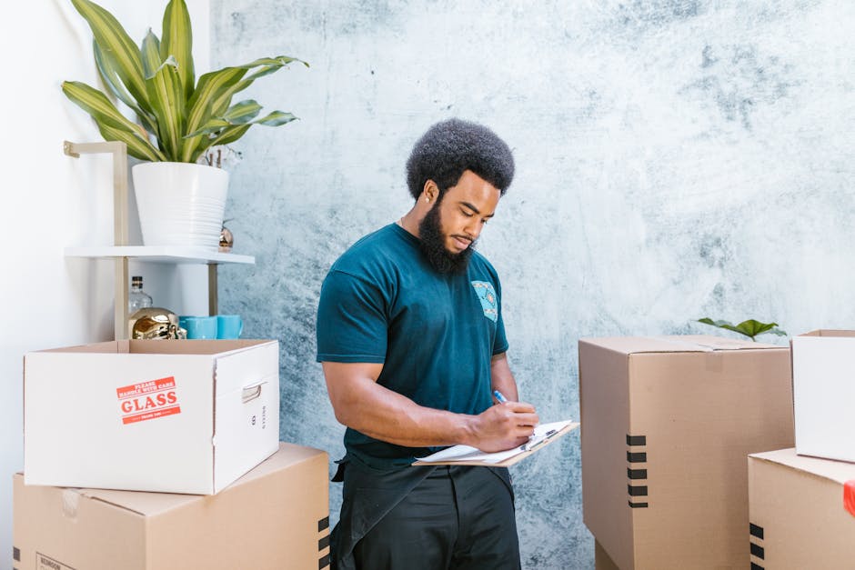 A professional mover from Man with Van Harefield is indoors, standing next to packed cardboard boxes during the home relocation process. He is wearing a blue company T-shirt and black trousers, holding a clipboard in one hand and writing with the other. The boxes are sealed with tape, some labeled with orange stickers indicating fragile items such as glass, and are stacked on each other and on the floor. Behind him, there is a white shelving unit against a textured white wall, holding a potted plant with large green leaves, decorative items, and small teal containers. The environment appears well-lit, indicating natural light or indoor lighting suitable for packing and moving activities. Visible equipment includes a trolley or hand truck in the background, assisting with the furniture transport and loading process, which are part of the services offered by Man with Van Harefield in the context of house removals or moving logistics, as referenced by the UB9 moving checklist page.